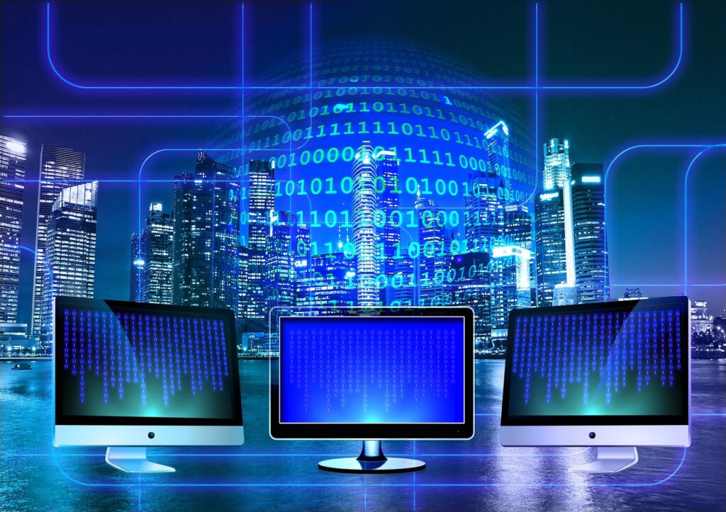 Three computer monitors displaying blue binary code in front of a brightly lit city skyline, representing digital technology and global connectivity.